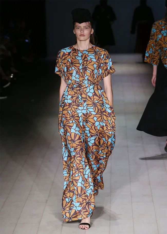 Model wearing a floral-patterned dress in shades of brown and blue on a runway.