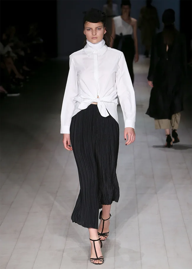 Model in a white blouse and black skirt walks runway, fashion show setting.