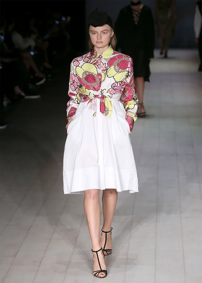 Model in white skirt and floral blouse on runway, wearing black sandals and hat.