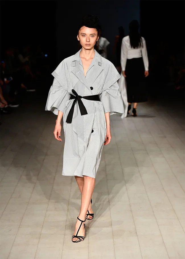 Model in black and white dress with belt walks runway; black heels and headpiece. White and black outfit in chic style.