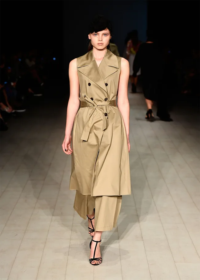 Model on runway wearing a beige sleeveless trench coat and wide-leg pants, with black strappy heels and headpiece.