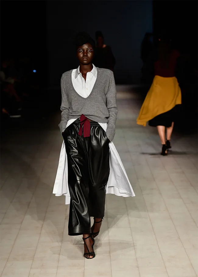 Model walks runway in layered fashion: gray sweater, white shirt, black wide-leg pants, burgundy and white accents.