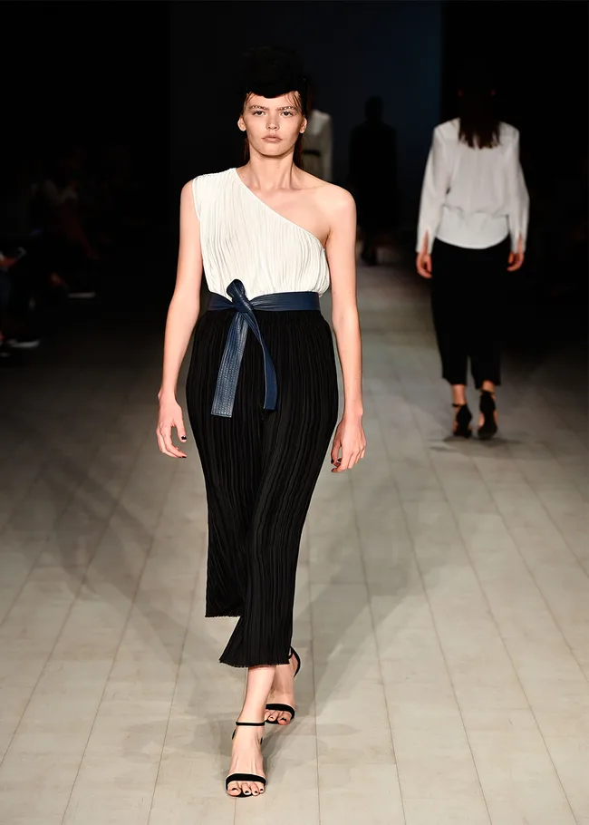 Model in a one-shoulder white top and pleated black skirt walks the runway with a confident expression.