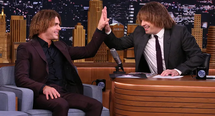 Two men with shaggy hairdo high-five on a talk show set, cityscape backdrop.