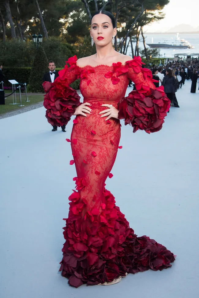 A woman in a glamorous red gown with floral details poses on a blue carpet, surrounded by trees and a distant yacht.