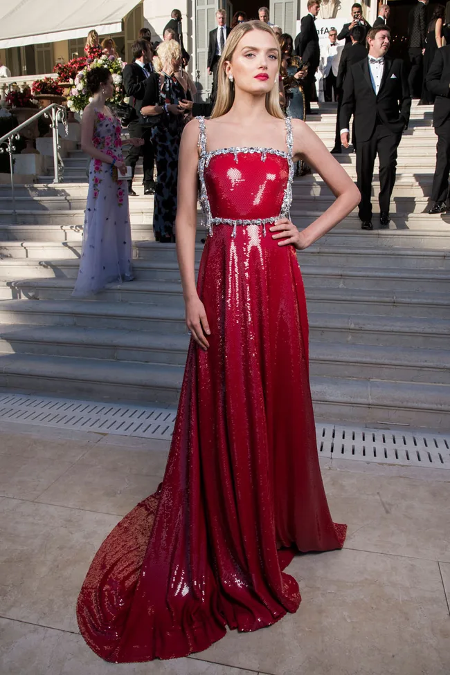 A woman in a shiny red gown poses on stairs at a formal event with people in the background.