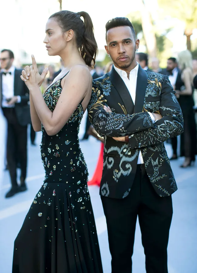 Two people in formal, embellished outfits, stand back-to-back at an outdoor event, one in a James Bond pose.
