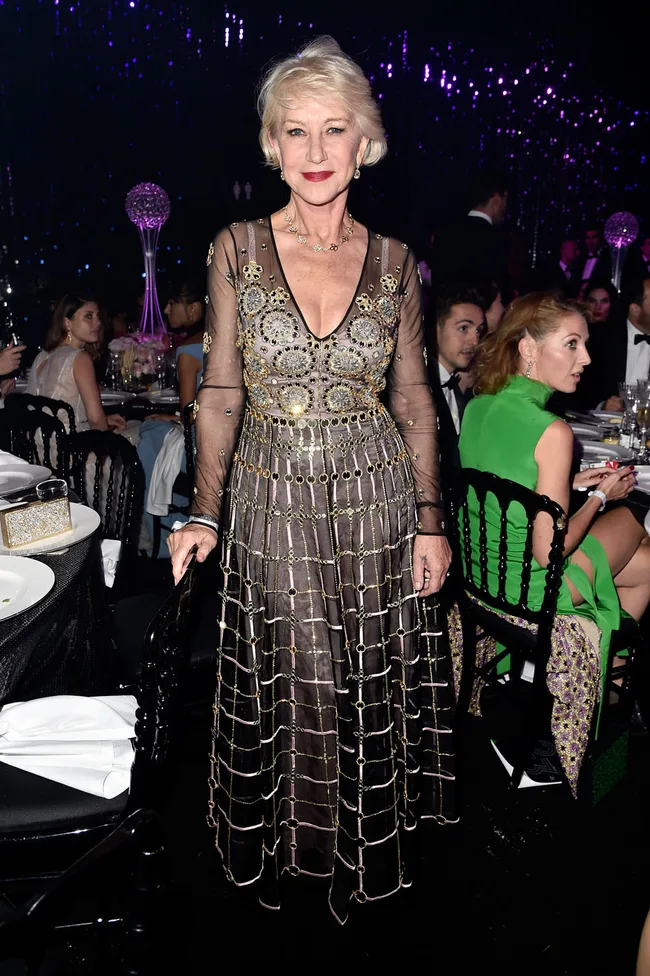 Elegant woman in a detailed gown, standing at a formal event, surrounded by seated guests and decorative lighting.