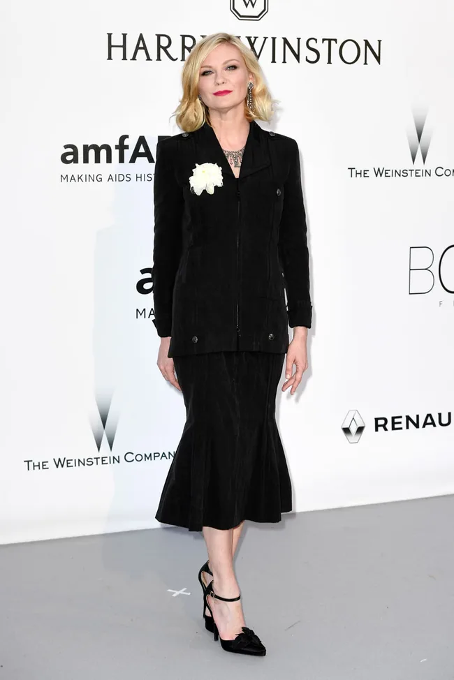 A woman in a black suit with a white flower, posing at an event with brand logos in the background.