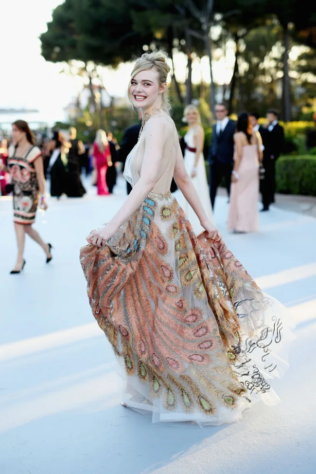 A person in a colorful, flowing gown smiles at an outdoor event, with people and trees in the background.