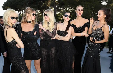 Group of six women in elegant black outfits, wearing sunglasses, outdoors at an event.