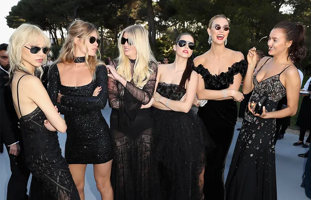 Group of six women in elegant black outfits, wearing sunglasses, outdoors at an event.