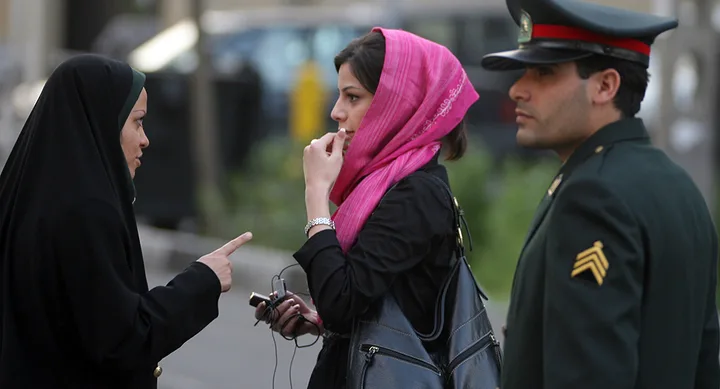 A woman in black chador talks to a woman in a pink headscarf, with a uniformed officer standing nearby.
