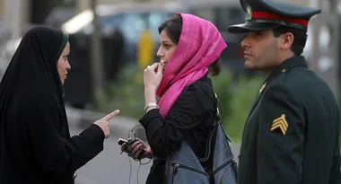 A woman in black chador talks to a woman in a pink headscarf, with a uniformed officer standing nearby.
