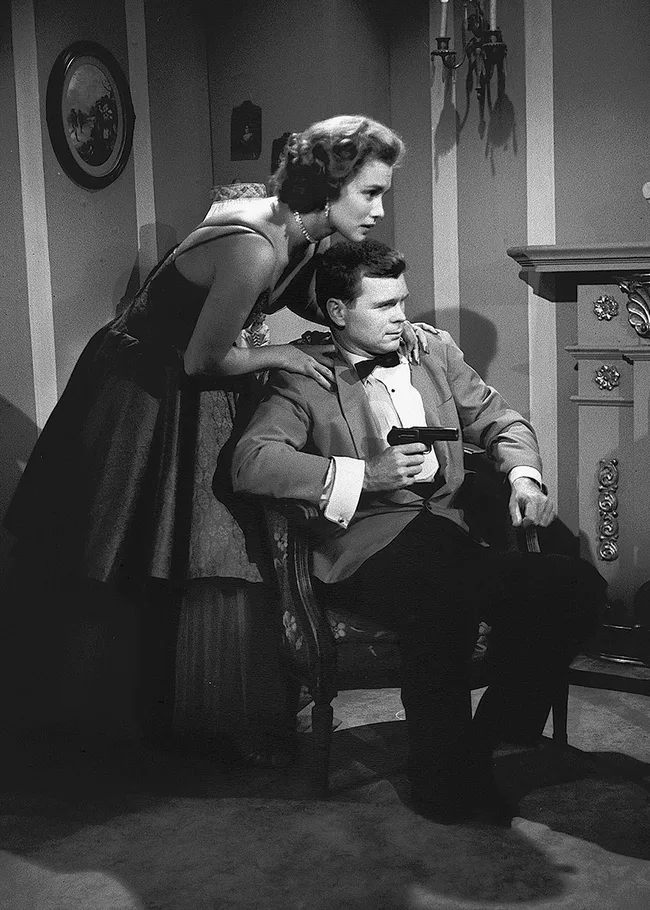 A woman leans over a seated man holding a gun, both in formal attire, in a vintage black-and-white setting.