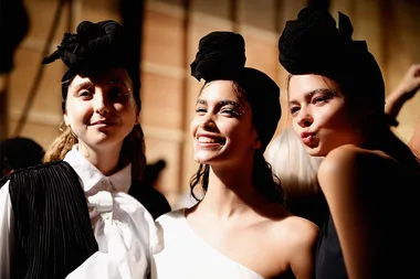Models in stylish outfits and headwraps smile and pose backstage at a fashion show.