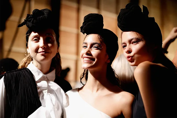 Models in stylish outfits and headwraps smile and pose backstage at a fashion show.