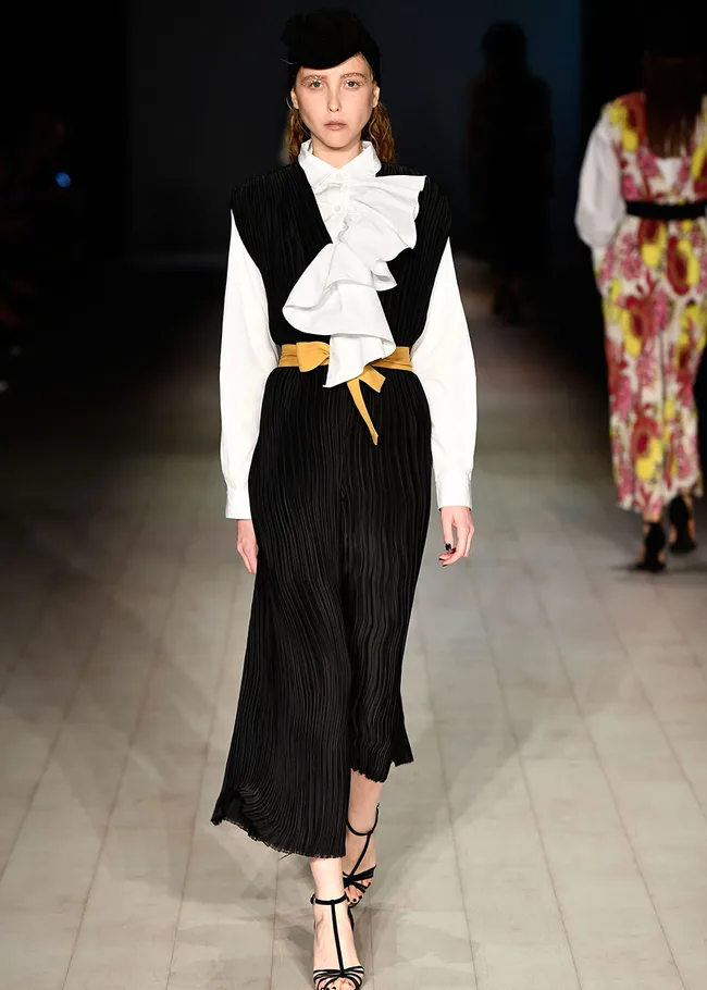 Model wearing a black pleated dress with a white ruffled top and gold belt on a runway.