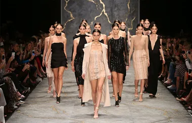 Models walk the runway in a fashion show, wearing diverse, elegant dresses, with a marble-patterned backdrop.