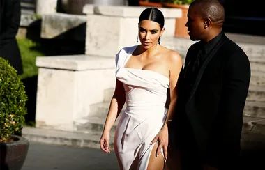 A woman in a white dress and a man in a black suit walk outdoors, with stone steps and greenery in the background.