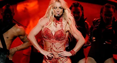 A performer in a glittering red outfit sings and dances energetically on stage with backup dancers.