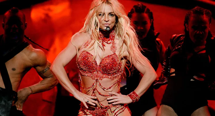 A performer in a glittering red outfit sings and dances energetically on stage with backup dancers.