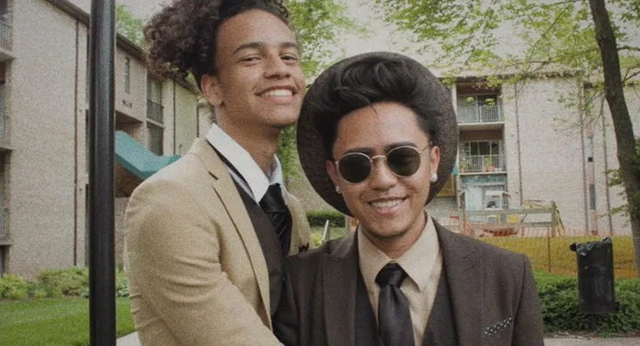 Two people smiling in suits, one wearing sunglasses and a hat, standing outdoors with trees and buildings in the background.