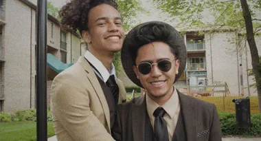 Two people smiling in suits, one wearing sunglasses and a hat, standing outdoors with trees and buildings in the background.