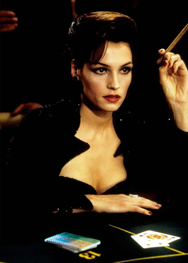 A woman holding a cigarette, dressed in black at a casino table with a deck of cards in front of her.