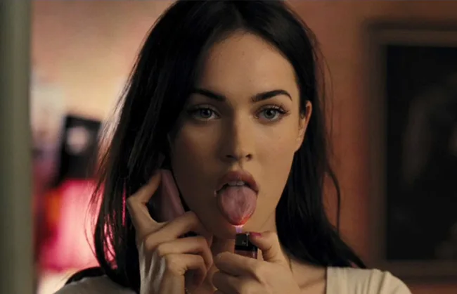 A person holds a phone and a lit lighter under their tongue, in a dimly lit room.