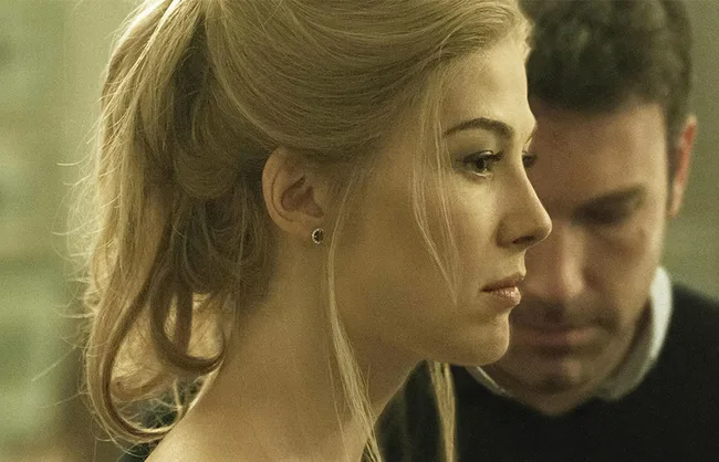 A blonde woman looks serious, with a man in the background appearing contemplative. The setting is subtly lit, adding dramatic tension.