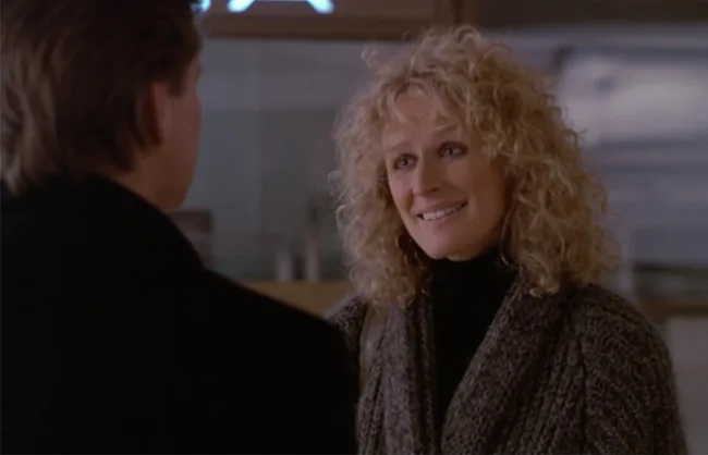 A woman with curly hair smiles at a man in an indoor setting, wearing a knitted sweater.