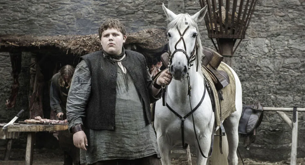 A person stands beside a white horse in a medieval setting with stone walls and a table with meat in the background.