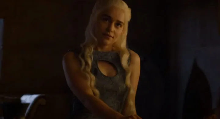 Blonde woman in a medieval-style setting, wearing a blue dress with a cutout, sitting and looking to her right.