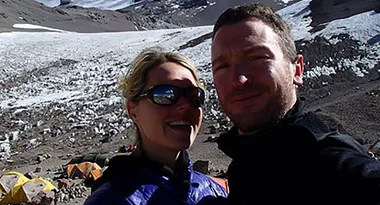 Couple taking a selfie on a snowy mountain with tents in the background, wearing outdoor jackets and sunglasses.