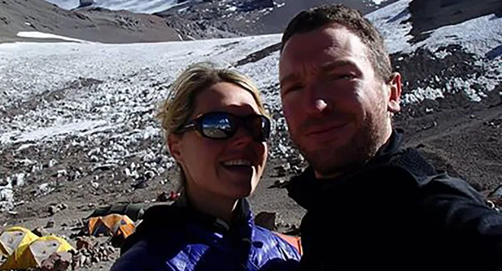 Couple taking a selfie on a snowy mountain with tents in the background, wearing outdoor jackets and sunglasses.