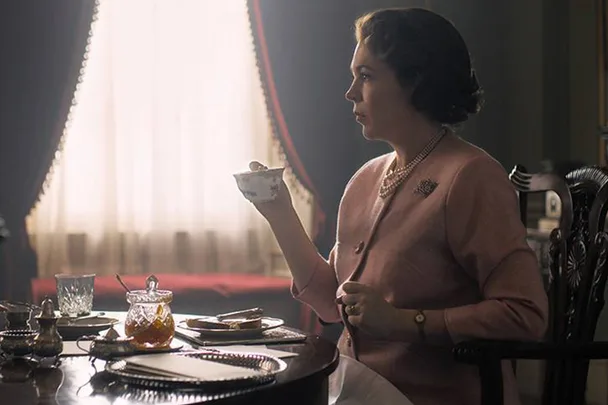 A woman in a pink suit sits at a table holding a teacup, with various items on the table and sunlight through a window behind her.