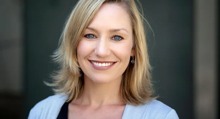 Blonde woman smiling with straight hair, wearing a light blue top. Neutral background.
