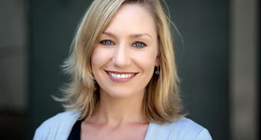Blonde woman smiling with straight hair, wearing a light blue top. Neutral background.