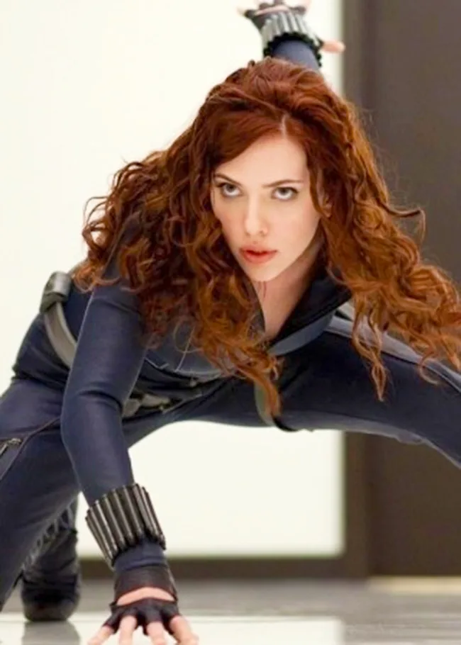 A woman in a black jumpsuit with curly red hair crouches on a glossy floor, ready to leap into action.