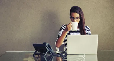 Woman in striped shirt using a laptop and drinking from a mug, sitting at a desk with a phone.