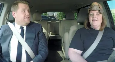 Two people sitting in a car, one wearing a black shirt and the other in a suit with a white shirt and tie, both smiling.