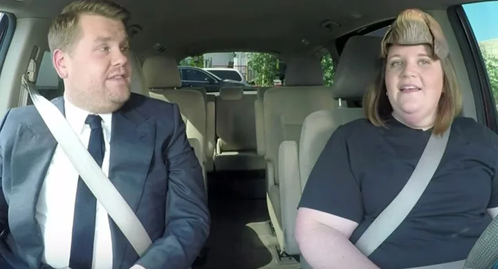 Two people sitting in a car, one wearing a black shirt and the other in a suit with a white shirt and tie, both smiling.
