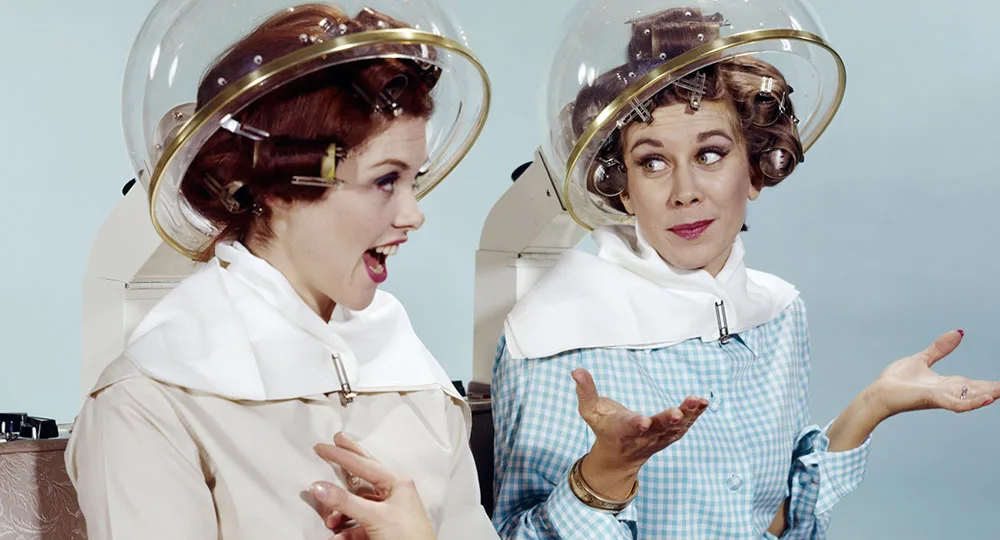 Two women with curlers under vintage hair dryers, chatting and gesturing, in a salon setting.