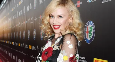 Blonde woman in floral dress smiles on red carpet with Alfa Romeo logo in background.