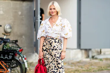 A woman in a puff-sleeve blouse with horse print and floral skirt holds a red bag, standing near a bicycle.