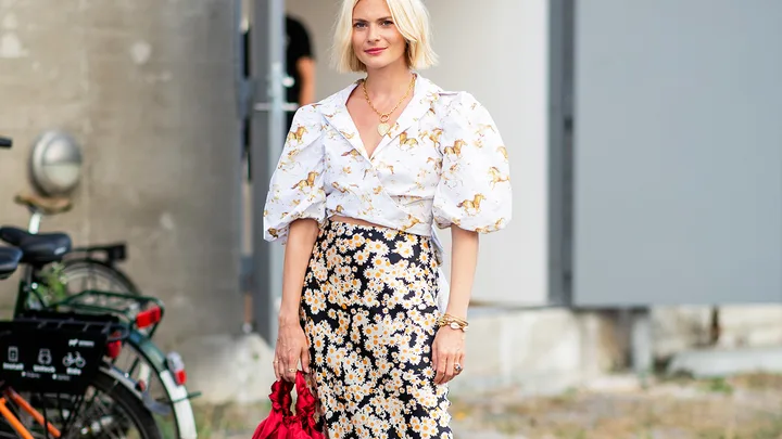 A woman in a puff-sleeve blouse with horse print and floral skirt holds a red bag, standing near a bicycle.