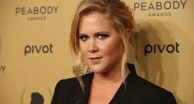 A person at the Peabody Awards wearing a black outfit against a gold background with text logos.