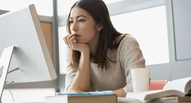 A woman in a sweater sits at a desk, concentrating on a computer screen, next to books and a coffee cup.