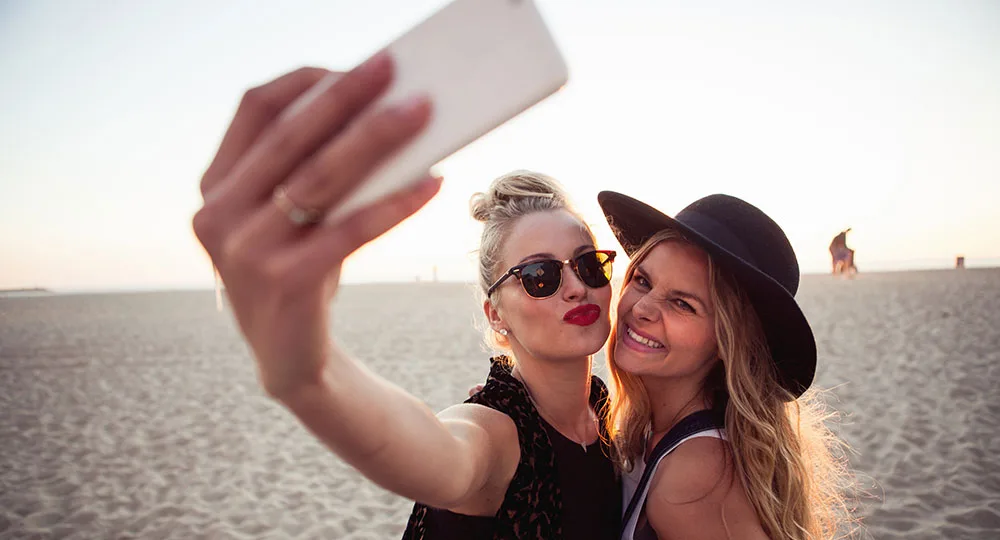 Two women on a beach taking a selfie; one wearing sunglasses and red lipstick, the other in a black hat, smiling.